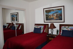 A hotel room with two beds, each with red bedding and matching pillows. There is a framed picture on the wall above the beds, and a small table with a lamp and a plant on the side.
