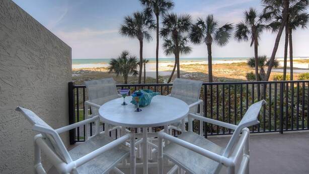 Een balkon met een witte tafel en stoelen, met uitzicht op een strand met palmbomen en een heldere lucht.