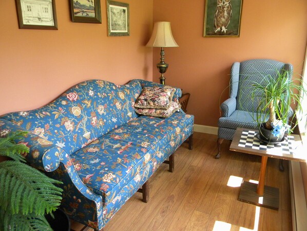 A living room with a blue floral sofa, a patterned armchair, a lamp, a potted plant, and framed pictures on the wall.