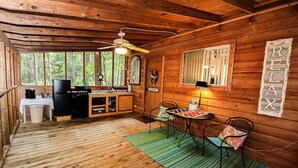 Interior rústico de una cabaña con piso de madera, un comedor con mesa y sillas, una cocina con refrigerador y pileta, y un amplio ventanal con vistas al bosque.