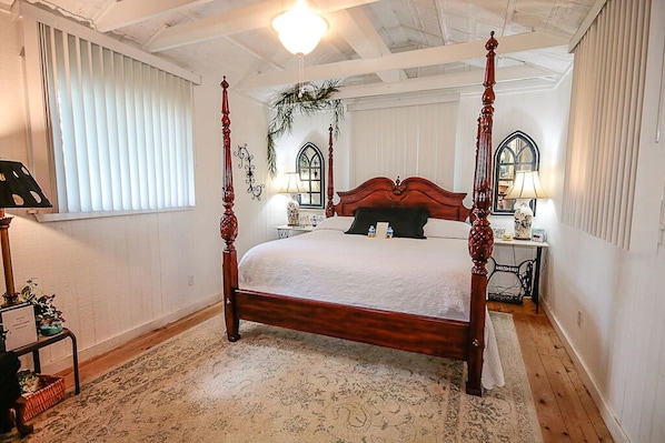 Uma cama de dossel com estrutura de madeira e roupa de cama branca, uma mesinha de cabeceira com abajur e uma janela com cortinas.