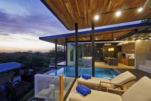 Un área de piscina moderna al aire libre con sillones de descanso y una cocina con paredes de vidrio.