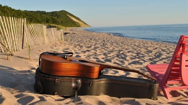 Une guitare et un verre sur une plage, avec une chaise rouge et une clôture en bois en arrière-plan.