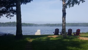 Une vue sur le lac, avec une jetée en bois qui s’avance dans l’eau, entourée d’arbres et agrémentée de trois chaises Adirondack.