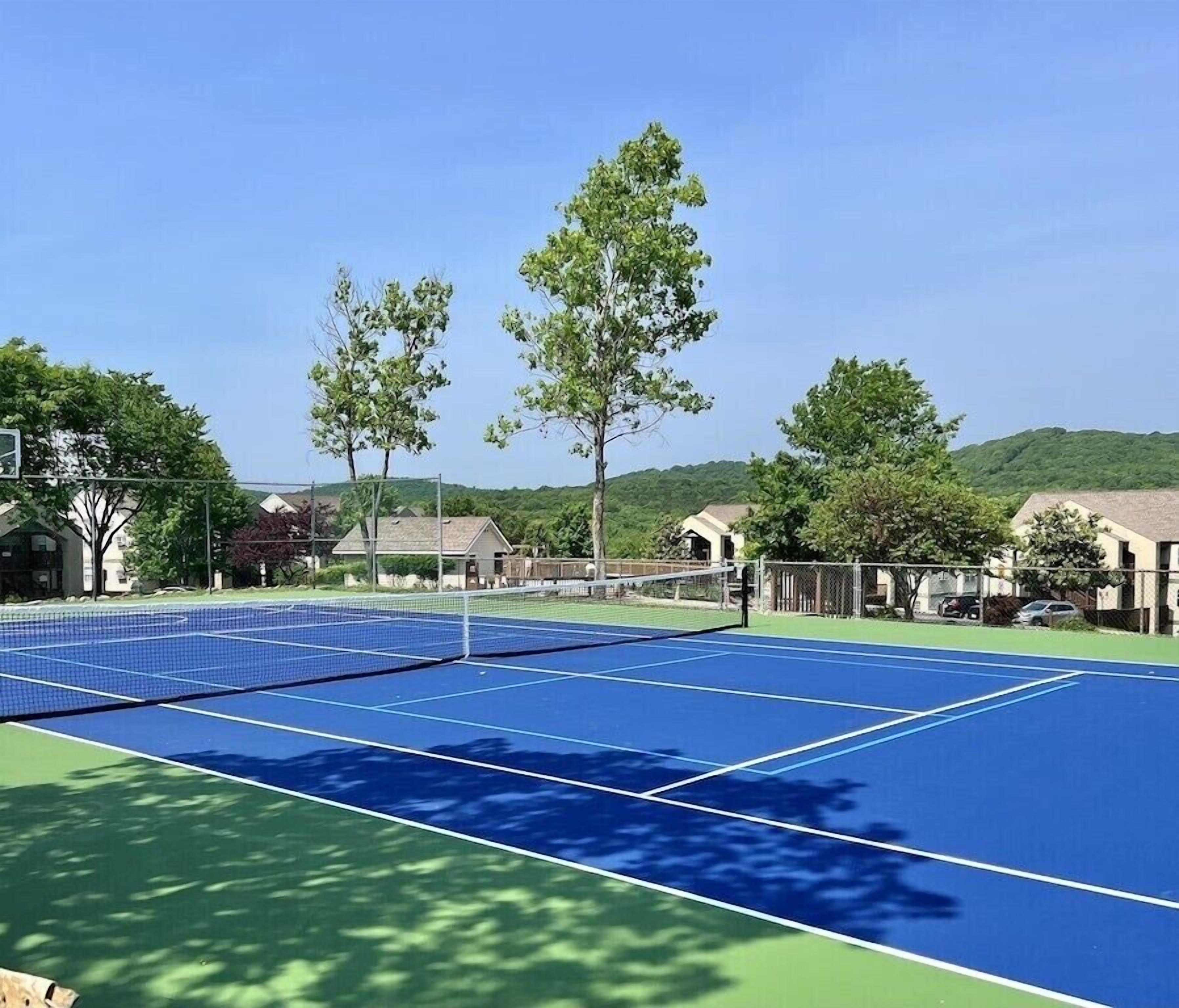 An outdoor tennis court with a net, surrounded by trees and houses.