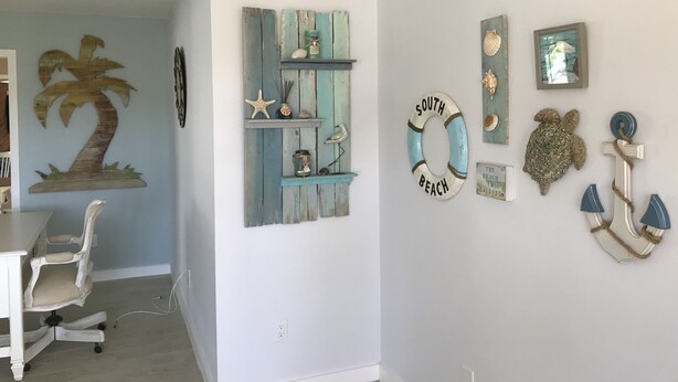 A room with a white desk, a white office chair, a wooden shelf with decorative items, and wall decorations including a palm tree, a lifebuoy, and a turtle. - Newly Renovated Coastal Style house (Sebastian)