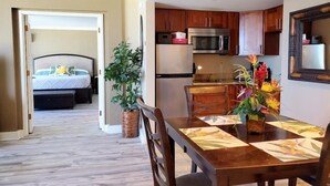 Habitación de hotel compacta con cama, zona de comedor con mesa y sillas, cocina con microondas y refrigerador, y una planta en maceta.
