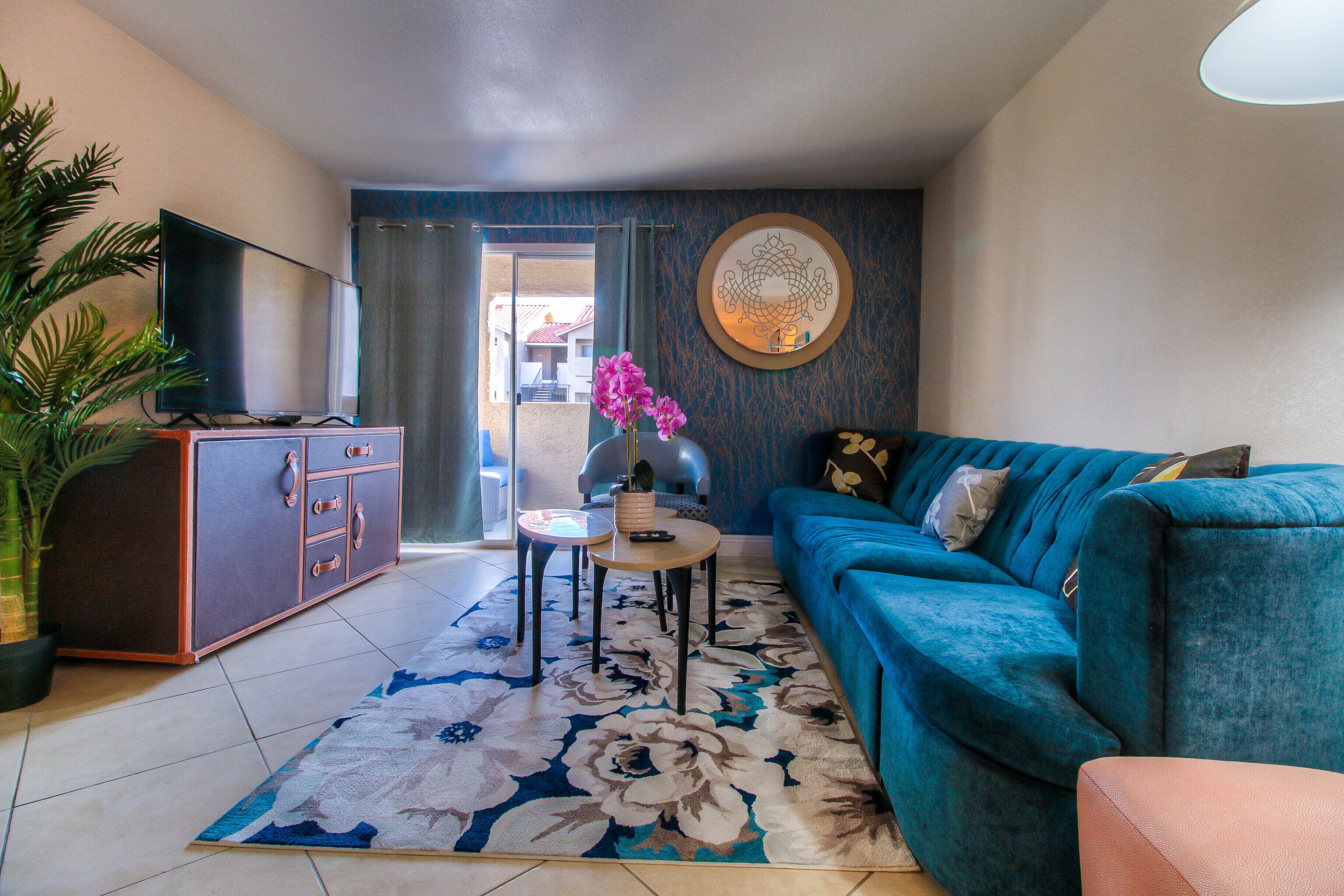 Una sala de estar con un sofá, un televisor sobre un mueble, una mesa de centro con un florero y una alfombra grande con estampado floral.