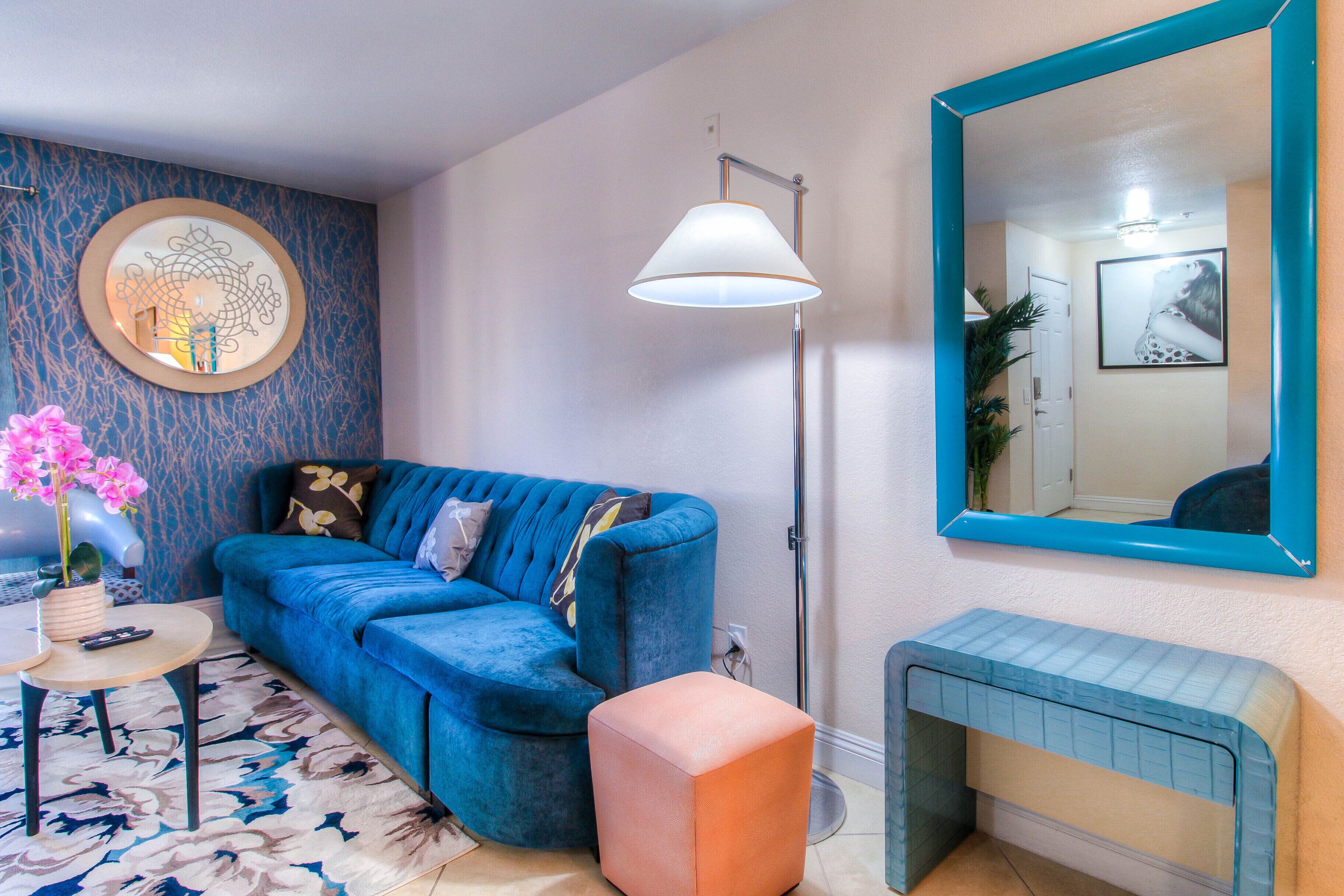 Ein modernes Wohnzimmer mit einem blauen Sofa, einem runden Spiegel, einer Stehlampe und einem kleinen orangefarbenen Ottomanen.