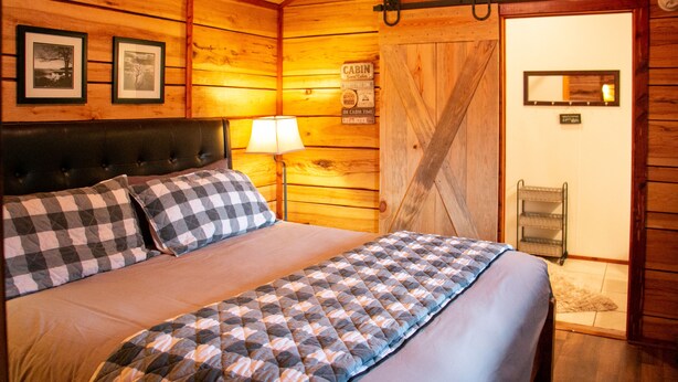 A cabin-style room with a bed, checkered pillows, a lamp, and a wooden door. - The Cabin at Panther Valley Ranch (Hot Springs)