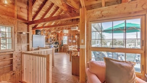 A wooden cabin interior with a dining area, kitchen, and a large window with an umbrella. - Sunset House: Rustic Maine Cottage Perched High Above Fishing Village with Harbor Views (Port Clyde)