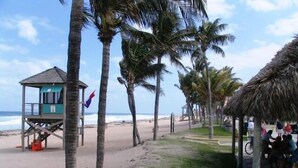 Une plage avec une tour de surveillance, des palmiers et une structure en chaume sous laquelle des personnes sont assises.