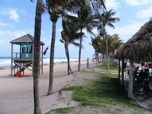 A beach with a lifeguard tower, palm trees, and a thatched structure with people sitting underneath. - Direct Oceanfront Luxury, Beautifully Redone! Satisfaction Guaranteed (Deerfield Beach)