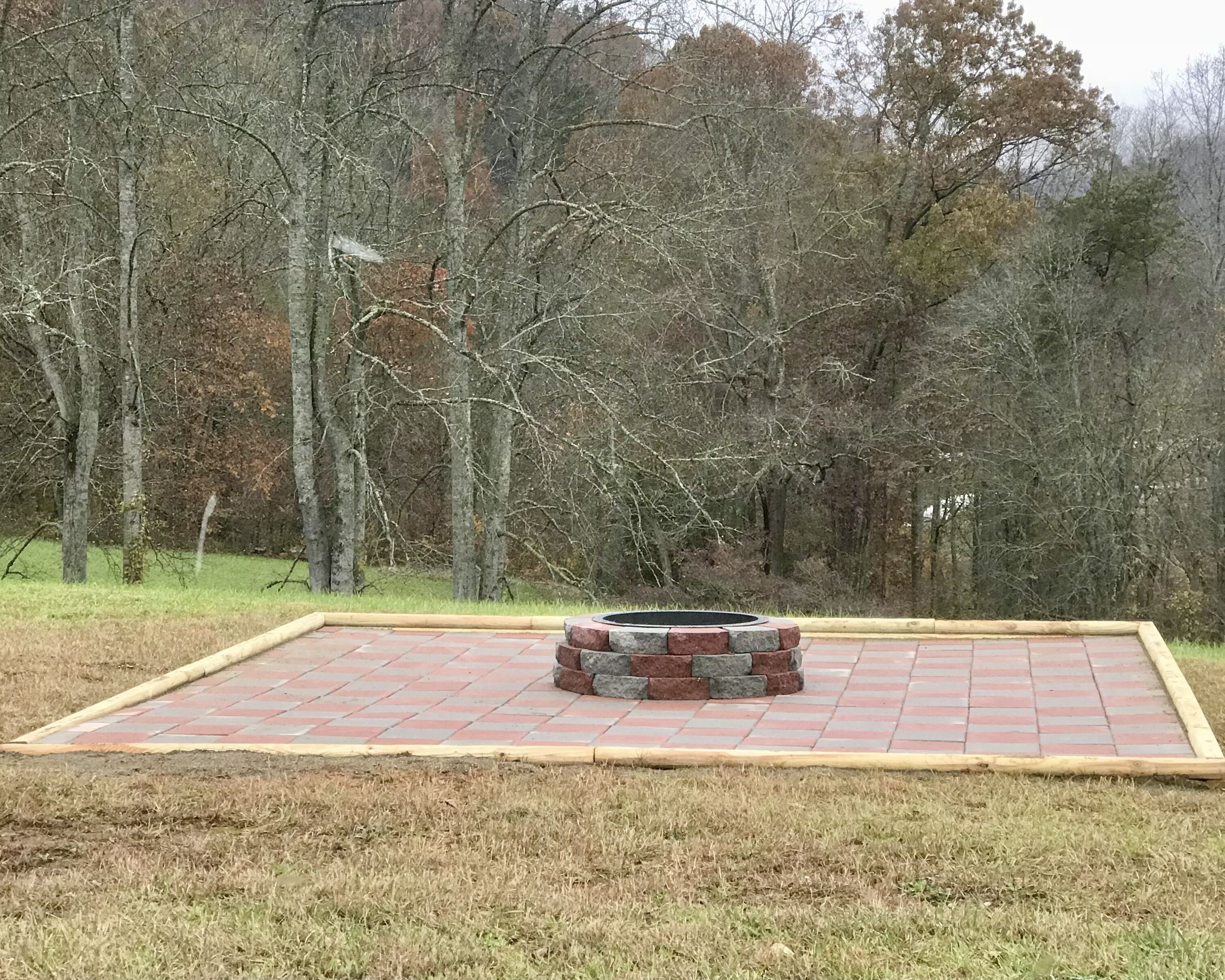 A fire pit surrounded by a brick border in a grassy area with trees in the background.