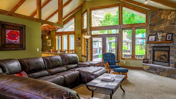 A spacious living room with a leather sofa, a stone fireplace, and large windows offering a view of greenery.