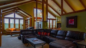 A spacious living room with a large sectional sofa, a coffee table, and a fireplace. There are large windows providing natural light and a view of the outdoors.