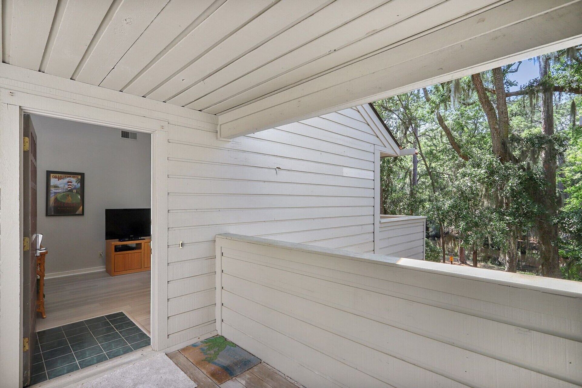 Eine überdachte Veranda mit Blick auf Bäume, eine Tür zu einem Raum mit Fernseher und ein kleiner Teppich.