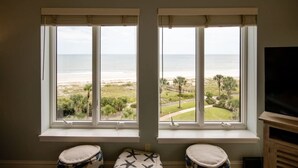 Une pièce avec deux fenêtres donnant sur une plage, un téléviseur sur le côté droit et une petite table avec un coussin.