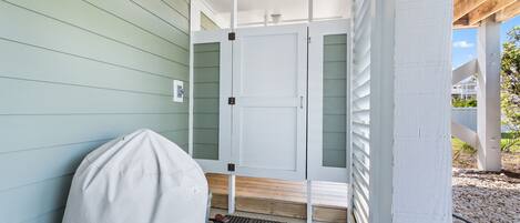 A covered porch with a white door, a covered grill, and a wooden bench.