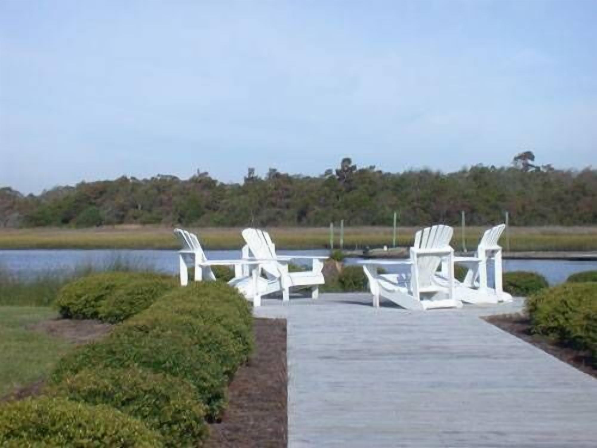 A wooden deck with white Adirondack chairs and a table, overlooking a body of water and a forested area.
