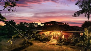 Ein traditionelles Haus mit Ziegeldach, umgeben von üppiger Vegetation und Palmen, unter einem leuchtenden Abendhimmel.