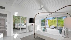A modern bedroom with a large bed, a ceiling fan, a TV, and a view of the outdoors.