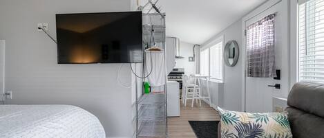 A modern hotel room with a bed, a sofa, a TV mounted on the wall, a kitchen area, and a bathroom with a shower curtain.