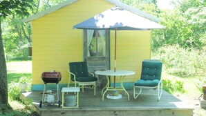 A yellow wooden structure with a porch, two green chairs, a small table, and a barbecue grill. - A cozy country "Doll House," with everything to meet your needs. (Hector)