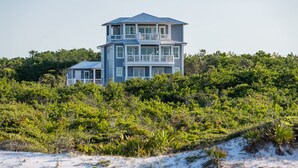 A two-story house with a balcony on the upper floor, surrounded by dense greenery and sandy terrain.