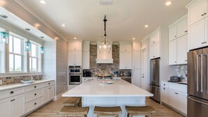 A spacious kitchen with white cabinetry, a central island with stools, stainless steel appliances, and a hanging light fixture.