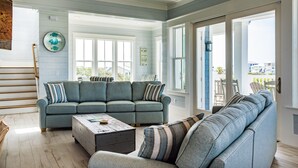 A spacious living room with a light blue color scheme, a large sectional sofa, a coffee table, and a view of the outdoors through large windows.
