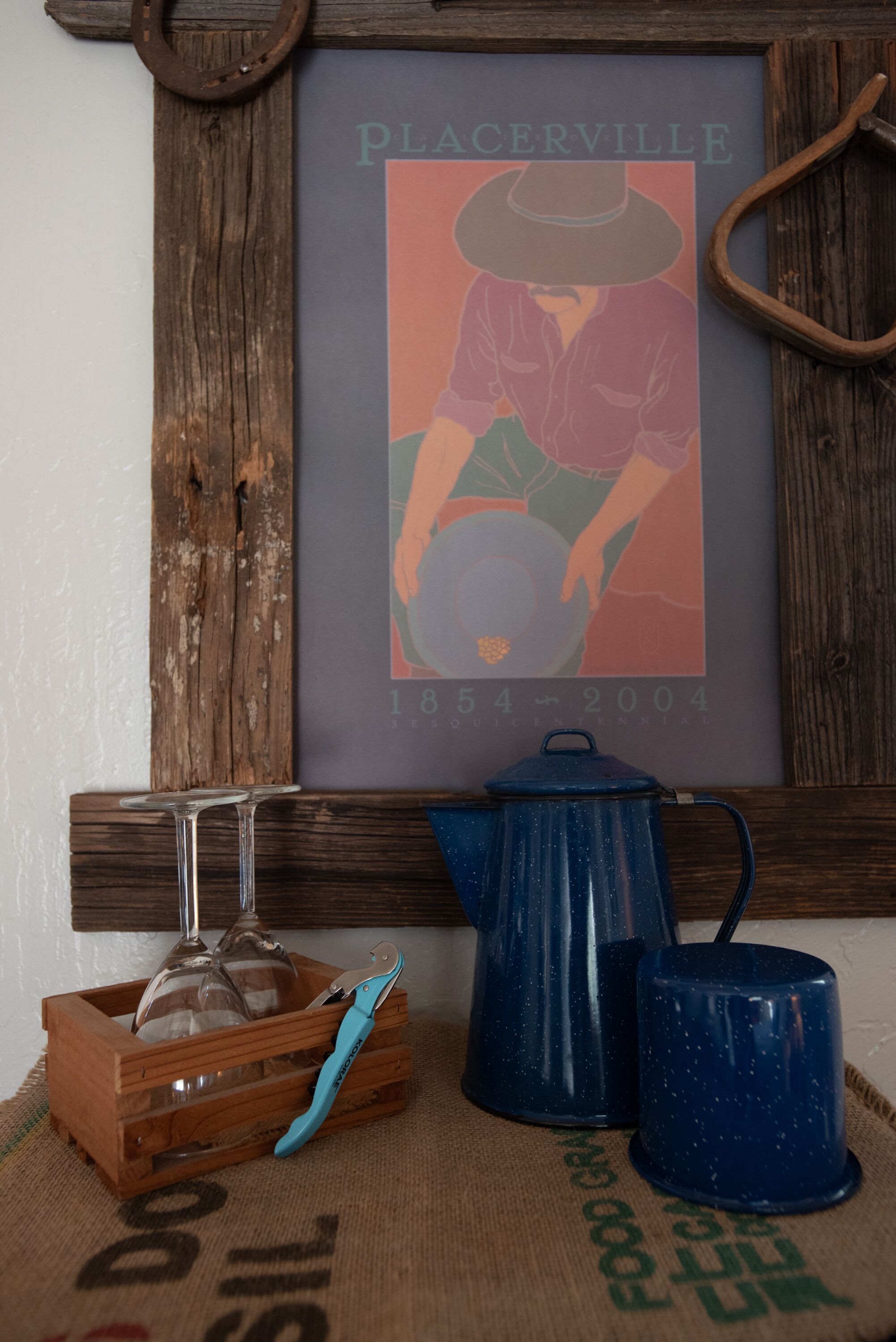 Una pared de madera rústica con un cartel enmarco de un hombre con sombrero, una tetera azul, un jarro de vidrio y una caja de madera con una herramienta de mango azul.