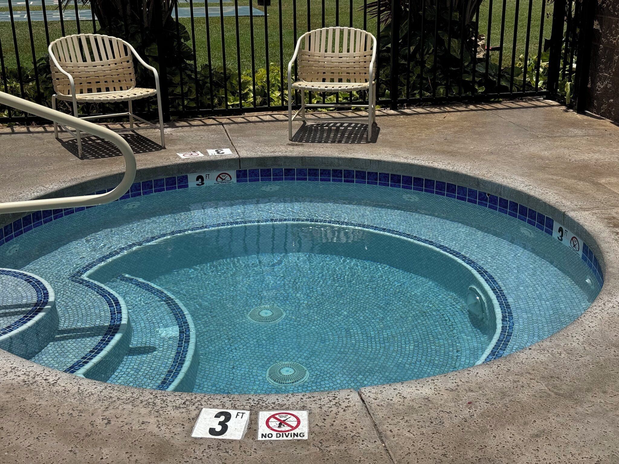 A small, circular pool with blue mosaic tiles, surrounded by a concrete deck. There are two white chairs and a metal handrail.
