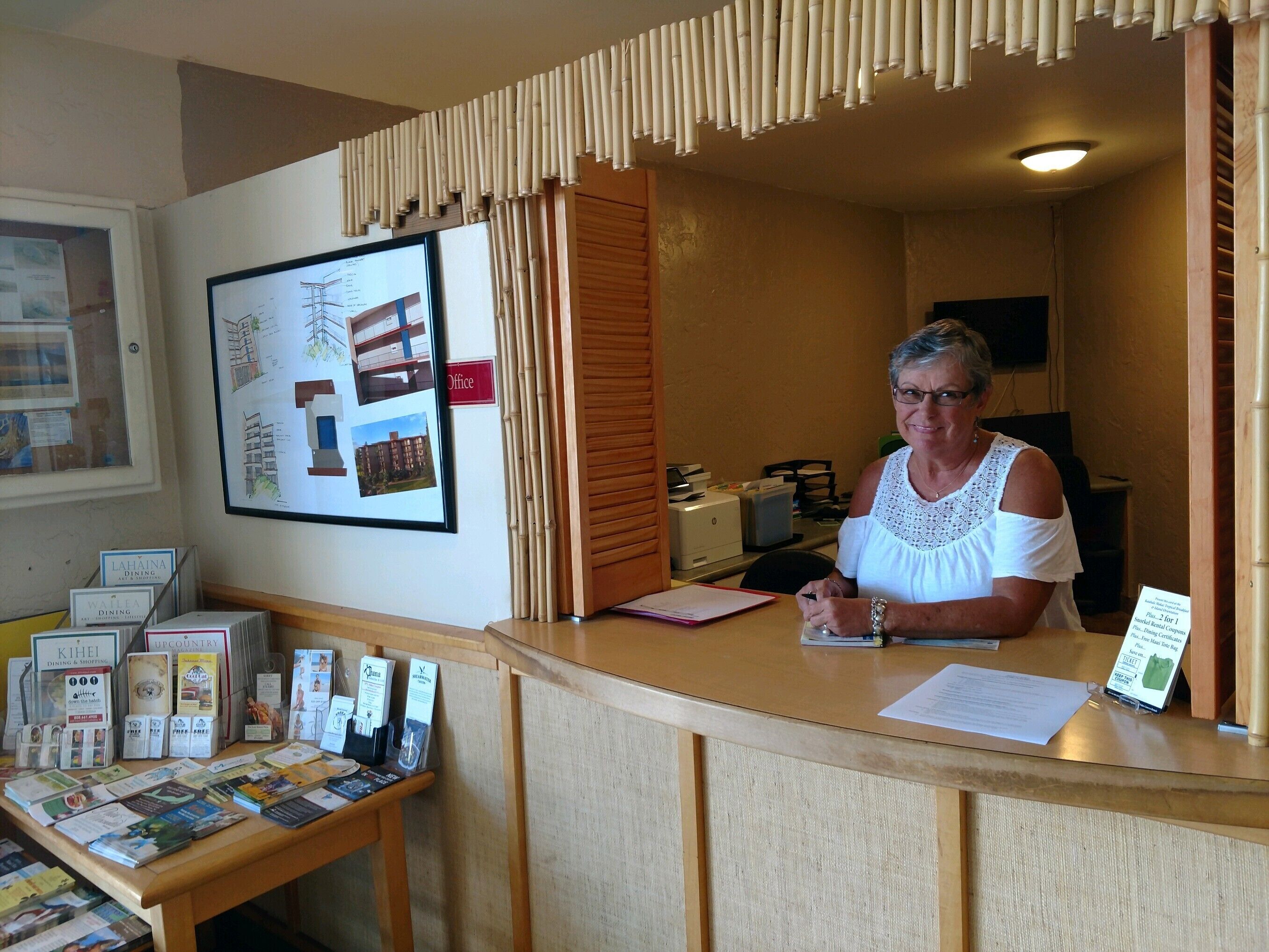 Una recepción con una recepcionista sentada, una pizarra con varios avisos y una exhibición de folletos y trípticos.