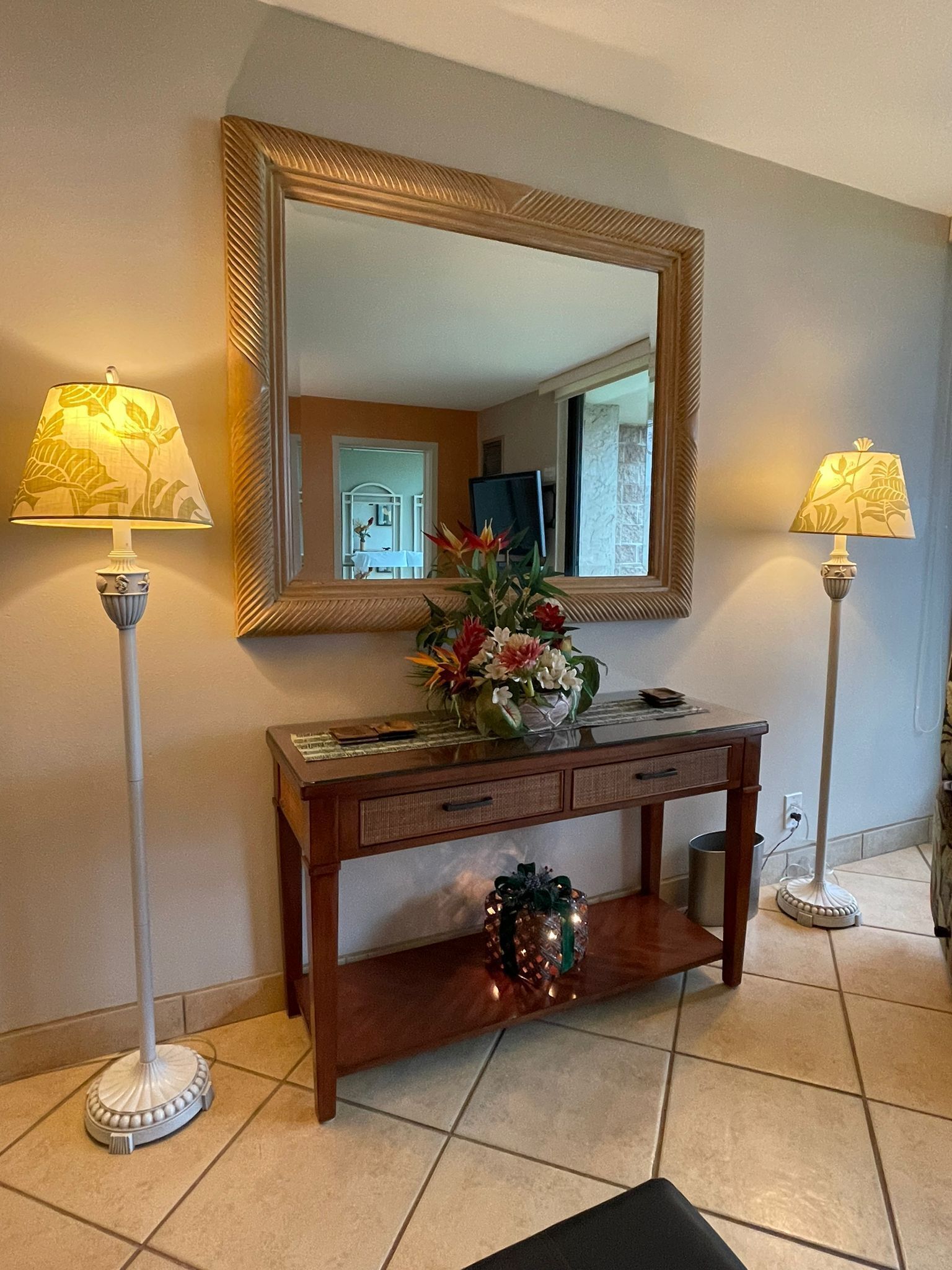 Een houten consoles tafel met een spiegel erboven, met aan weerszijden twee vloerlampen met lampenkappen, en een vaas met bloemen op de tafel.