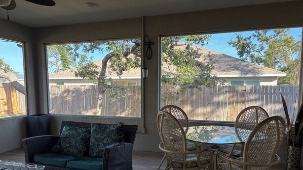 Une terrasse ensoleillée, meublée de meubles en osier, avec une table en verre et une vue sur une clôture en bois et des arbres.