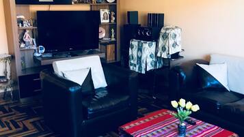 Una sala de estar con televisor de pantalla plana, sofás de cuero, una mesa de centro con camino de mesa colorido y una pequeña planta.