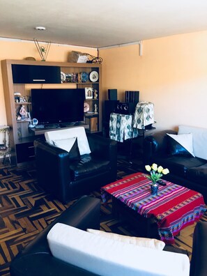Una sala de estar con televisor de pantalla plana, sofás de cuero, una mesa de centro con camino de mesa colorido y una pequeña planta.