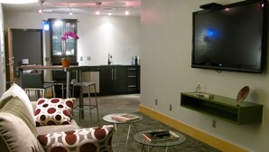 A living room with a sofa, a coffee table, and a TV mounted on the wall. There is a kitchen area in the background with a window, a plant, and a sink. - Suites at 118 (Bloomington)