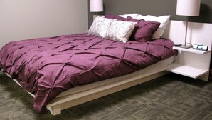 A neatly made bed with a purple bedspread and matching pillows, a white nightstand with a lamp, and a grey carpeted floor. - Suites at 118 (Bloomington)