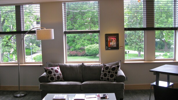 A living room with a gray sofa, a coffee table, and a floor lamp. There are large windows with blinds, a picture on the wall, and a view of greenery outside. - Suites at 118 (Bloomington)