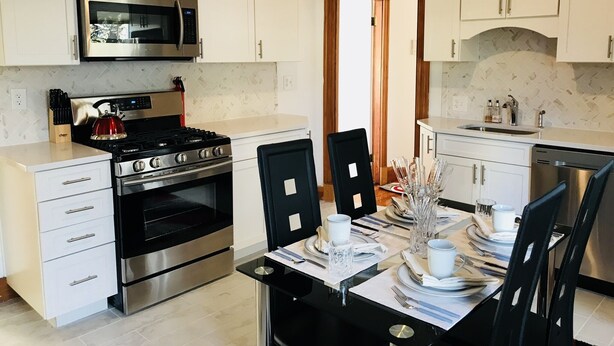 A modern kitchen with white cabinets, stainless steel appliances, and a dining area with a black table set for four. - Boston Luxury Suits Cottage for up to 24 guests!!! (Boston)