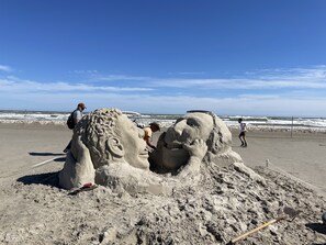 Une grande sculpture de sable représentant le visage d’un lion, sur une plage, avec un ciel dégagé.