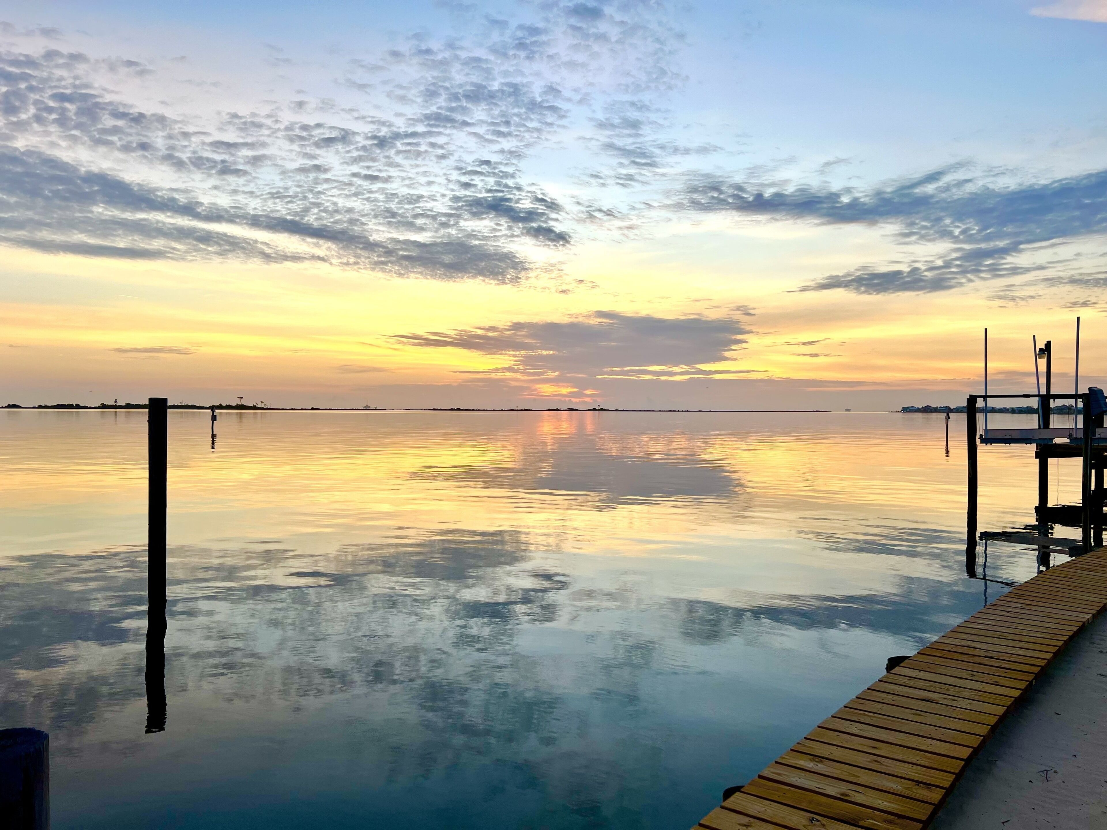 A calm waterfront view with a wooden dock extending into the water, a clear sky, and distant land.