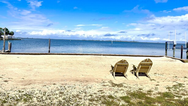 Ein Strand mit zwei Holzstühlen, die zum Wasser zeigen, einem Holzzaun und einem wolkenlosen Himmel.