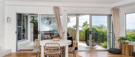 A modern living room with a dining area, large sliding glass doors leading to a balcony, and a view of greenery.