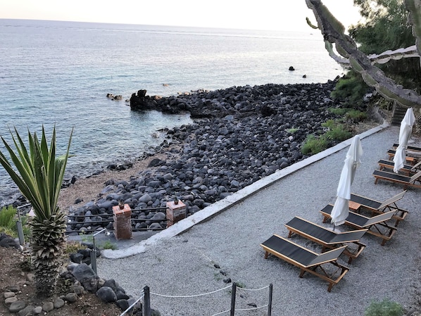 Zona costera con costa rocosa, sillas de descanso y una planta de palmera.