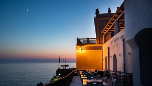 A waterfront restaurant with outdoor seating overlooking the sea, featuring a prominent entrance and a view of the water. - Hotel L’Ariana ISOLE EOLIE - UNA Esperienze (Leni)