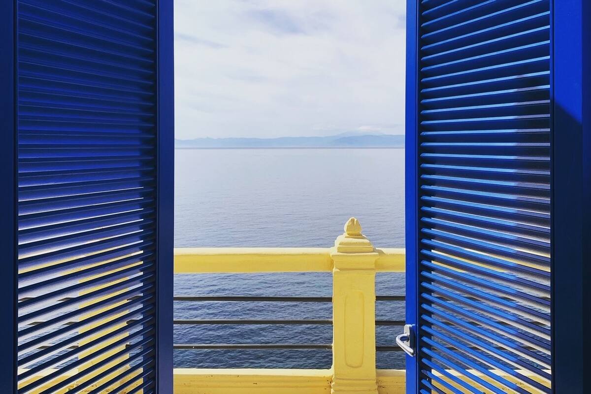 Blick durch blaue Fensterläden auf ein Gewässer mit einem entfernten Landteil und einem gelben Geländer.