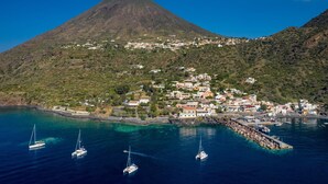 A coastal town with a mountain in the background, boats docked at a pier, and buildings along the shoreline. - Hotel L’Ariana ISOLE EOLIE - UNA Esperienze (Leni)